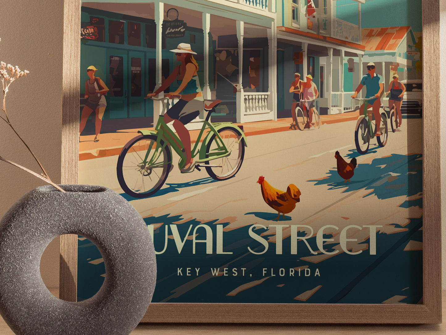 A colorful illustration of people biking on Duval Street, Key West, with roosters wandering freely. The framed artwork is set against a neutral wall with a decorative vase.