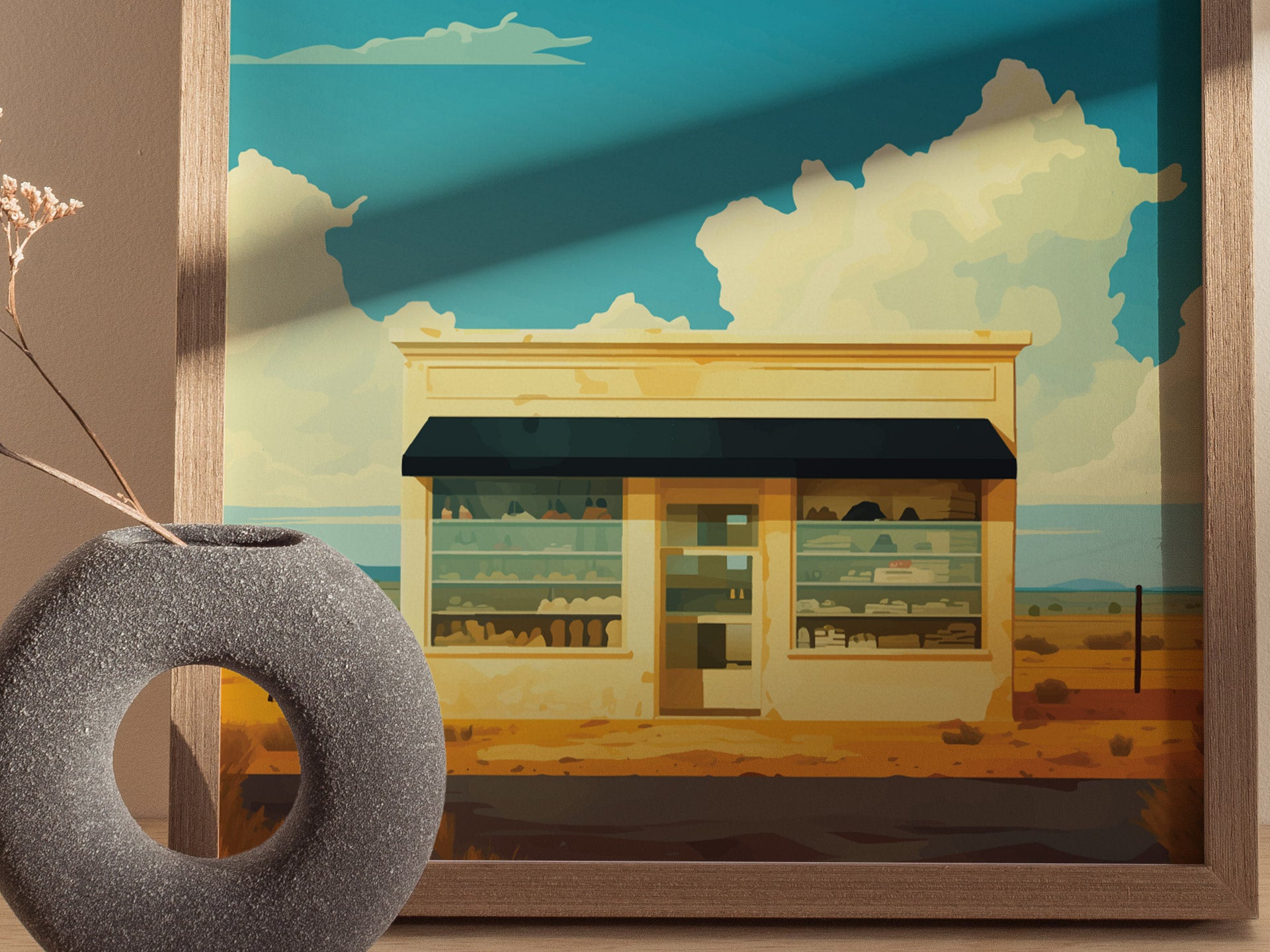 A framed poster depicts a desert art installation with a small building under a vast, cloud-filled sky. A textured vase with dried flowers is in the foreground.