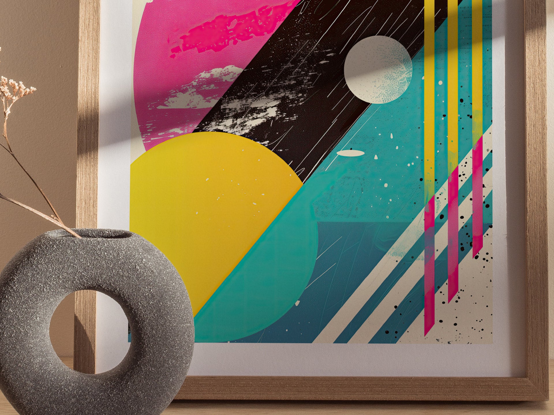 A framed abstract poster features bold geometric shapes in pink, yellow, and teal with distressed textures. A round, textured vase with a dried plant is in the foreground.