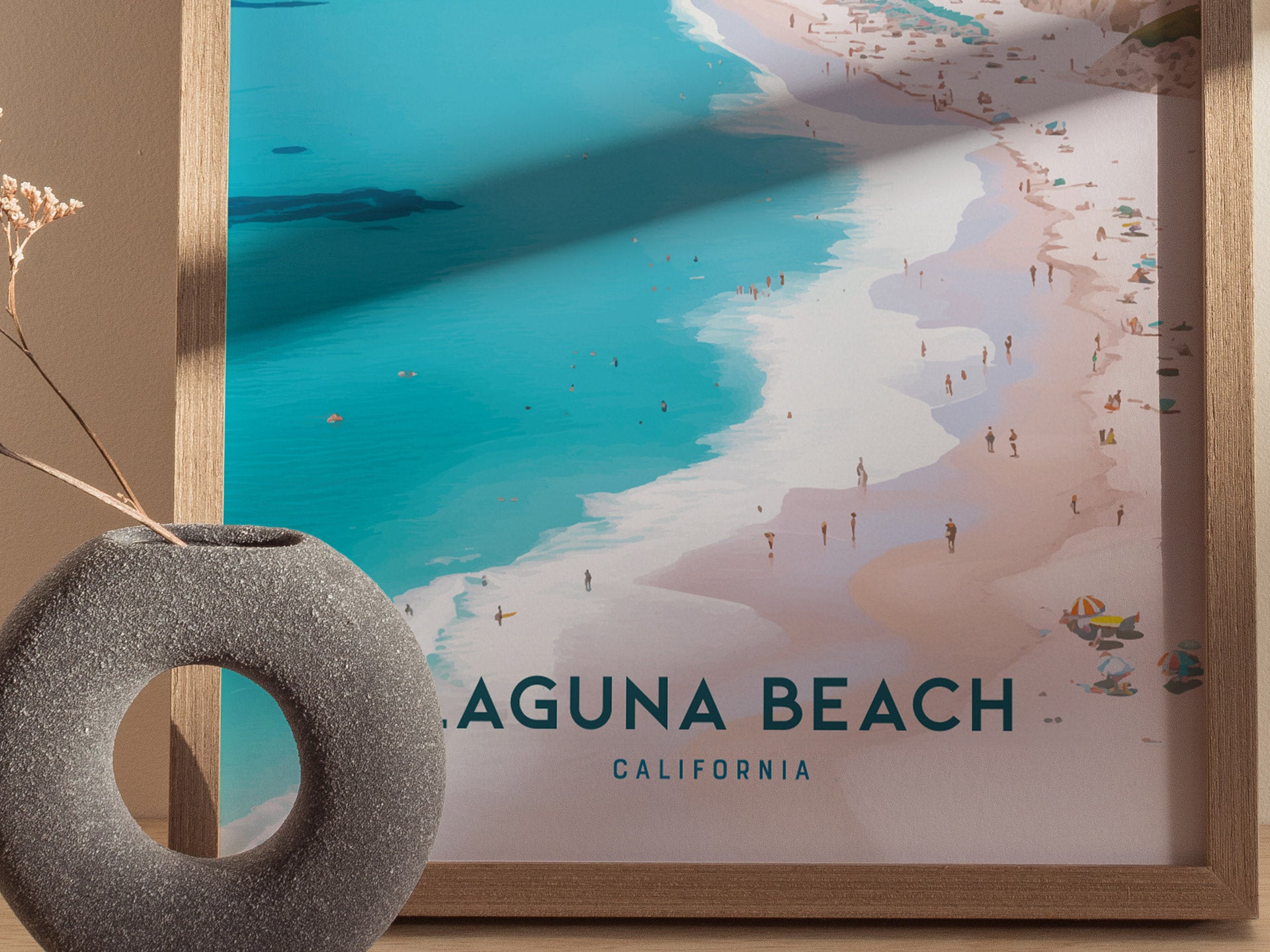 A framed Laguna Beach poster depicts a vibrant coastal scene with turquoise waters, sandy shores, and beachgoers. A stone vase with dried flowers is visible in the foreground.