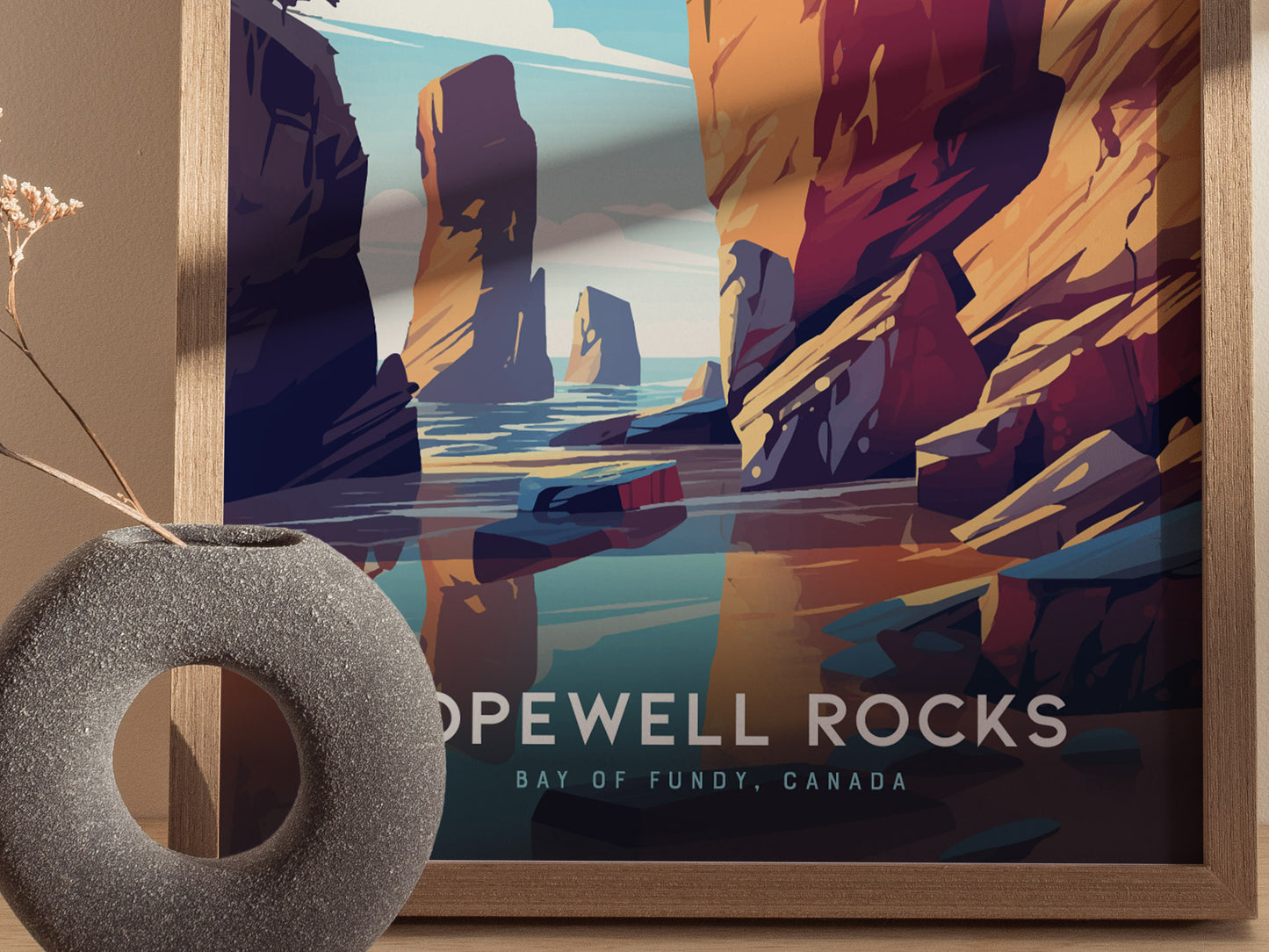 A framed poster of Hopewell Rocks in Bay of Fundy, Canada, featuring colorful cliffs and water reflections. A textured, circular vase with dried flowers sits in the foreground.