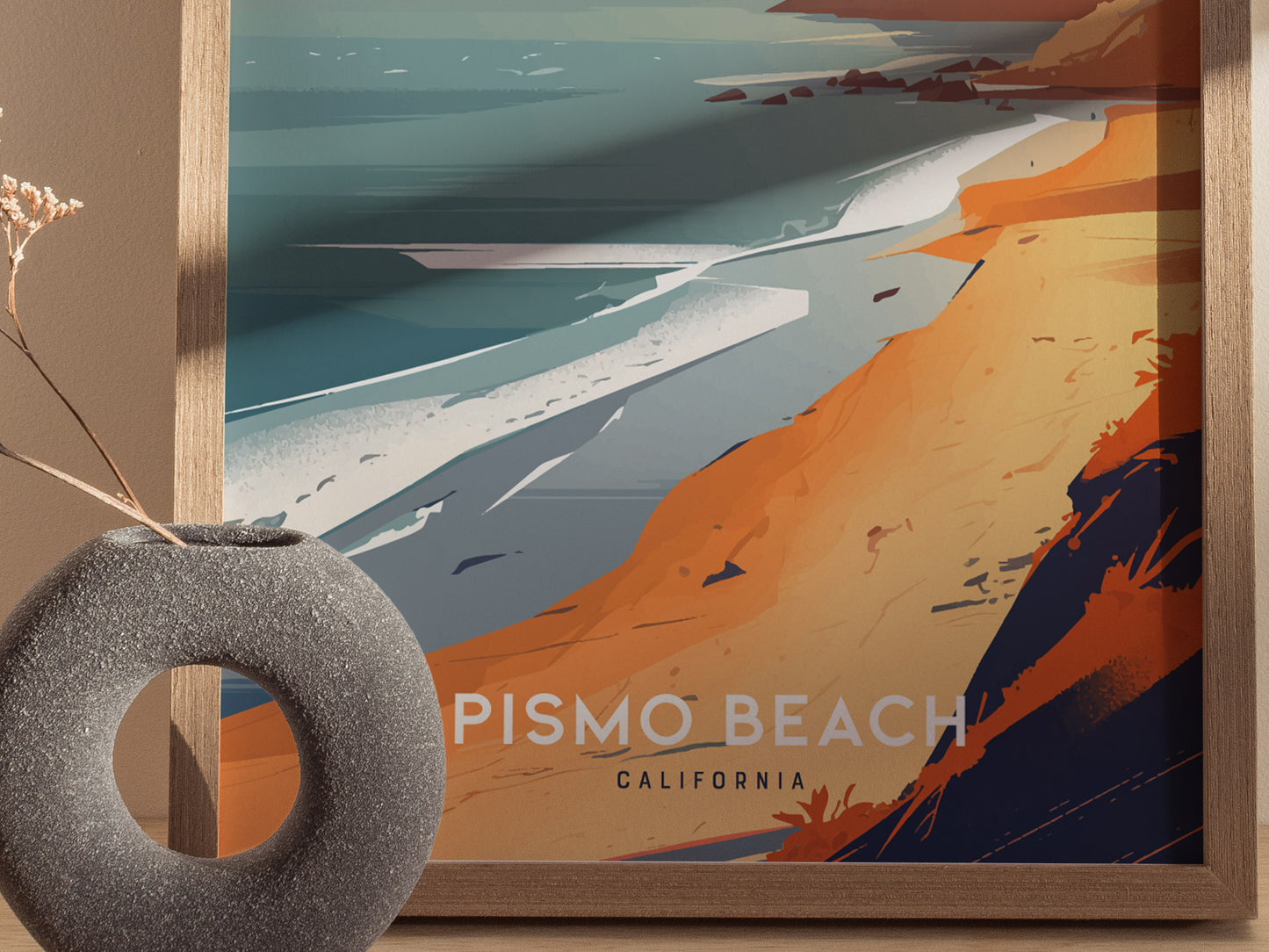 A framed poster depicts a stylized coastal scene with orange cliffs and blue ocean waves. The text Pismo Beach California is visible, with a gray vase in the foreground.