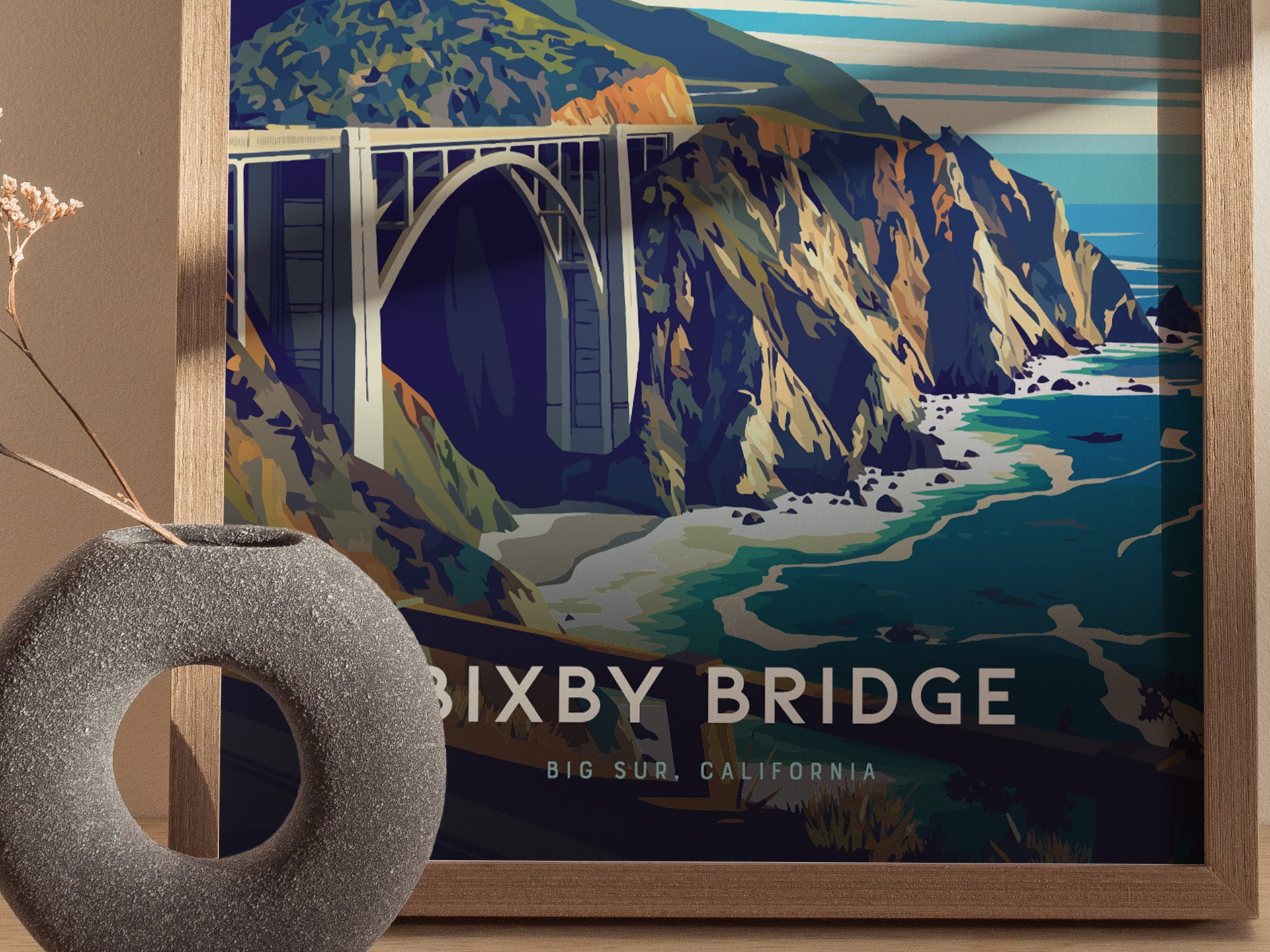 A framed poster depicts the Bixby Bridge spanning rocky cliffs and turquoise waters, with stylized mountains in the background. A textured gray vase with dried flowers sits nearby.
