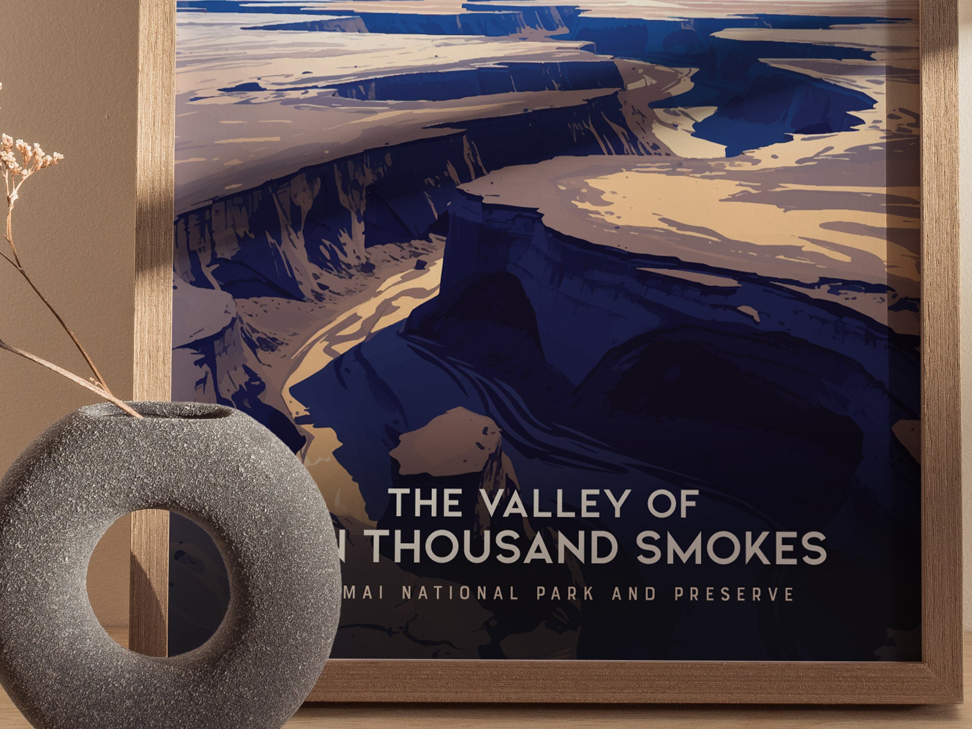 A framed poster of the Valley of Ten Thousand Smokes features a dramatic volcanic landscape with deep blue and beige tones. A round, textured vase with dried flowers is nearby.