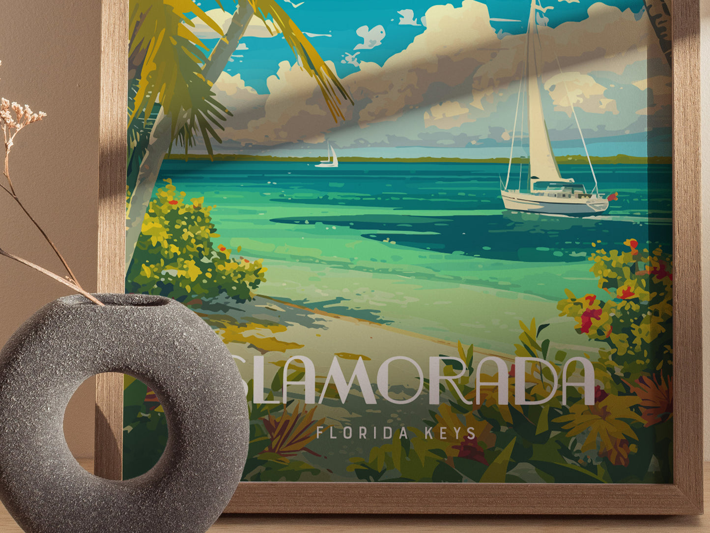 A framed poster of Islamorada, Florida Keys, features a sailboat on turquoise waters, tropical plants, and palm trees. A textured gray vase with dried flowers is in the foreground.