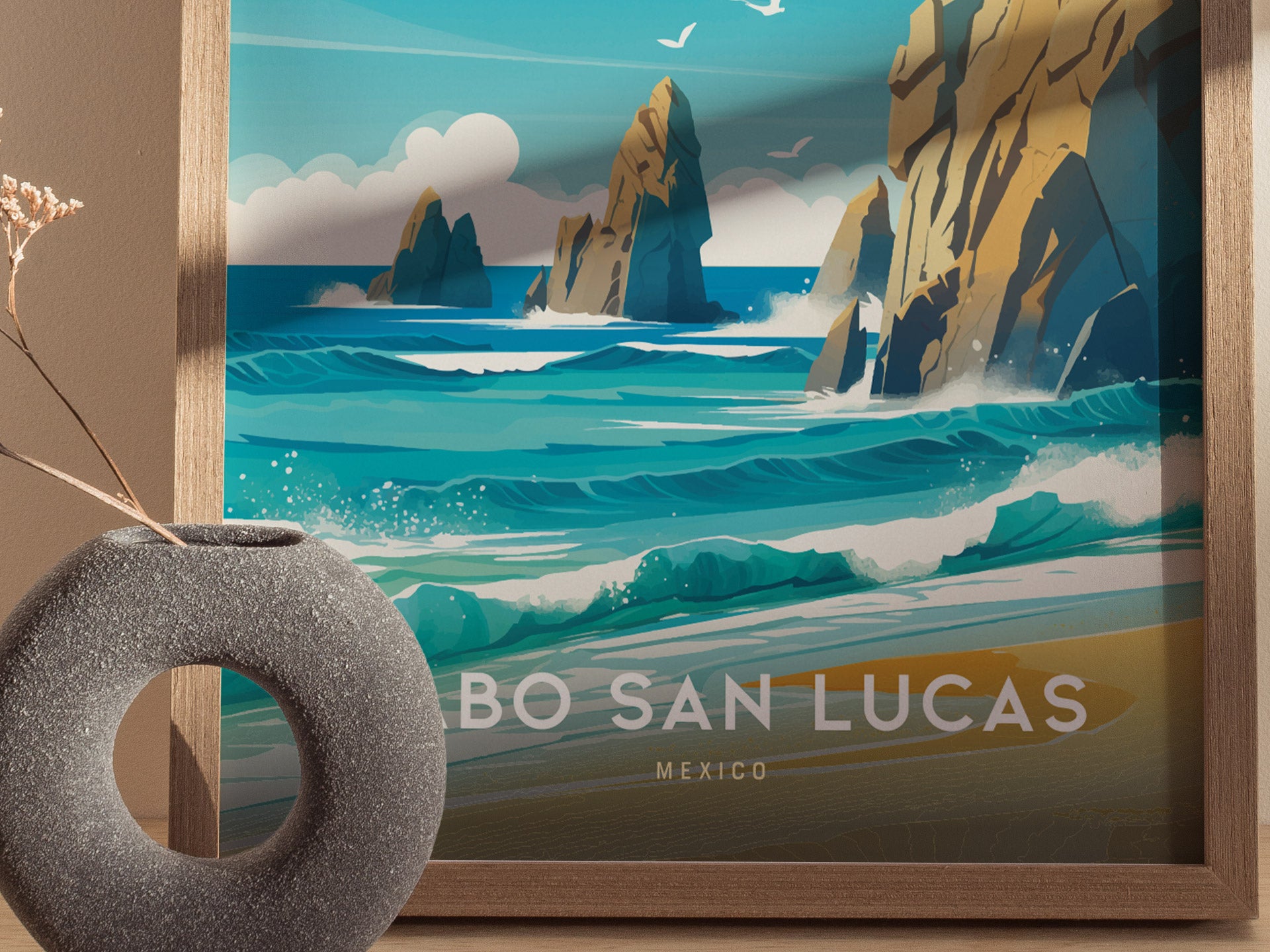 A framed Cabo San Lucas poster features turquoise waves crashing against rocky cliffs under a blue sky. A round, textured gray vase with dried flowers is placed in front.