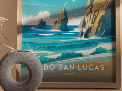 A framed Cabo San Lucas poster features turquoise waves crashing against rocky cliffs under a blue sky. A round, textured gray vase with dried flowers is placed in front.