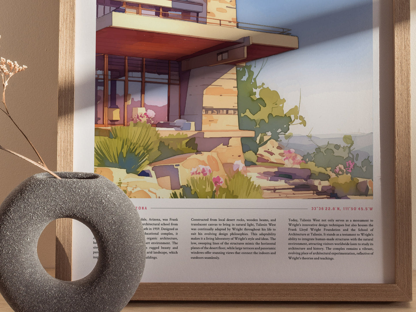 A framed poster of Taliesin West showcasing colorful desert architecture is displayed against a beige wall. A textured gray vase with dried flowers is positioned in the foreground.