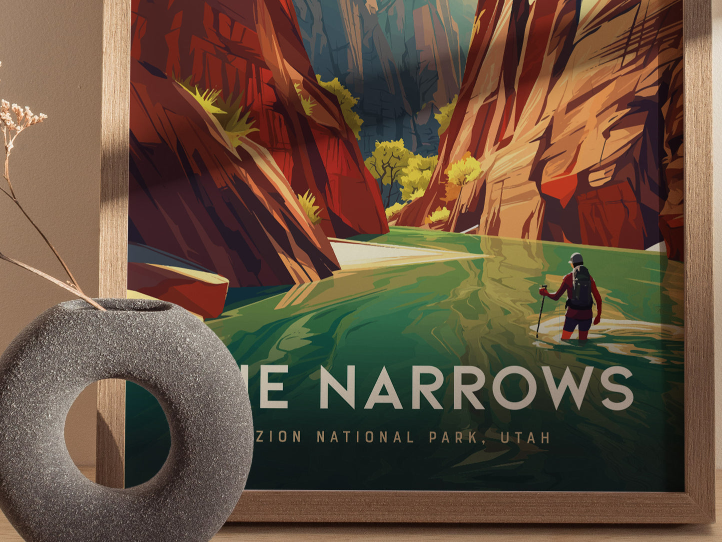 A framed poster of The Narrows in Zion National Park shows a hiker wading through a green river, surrounded by towering canyon walls. A round vase is placed nearby.