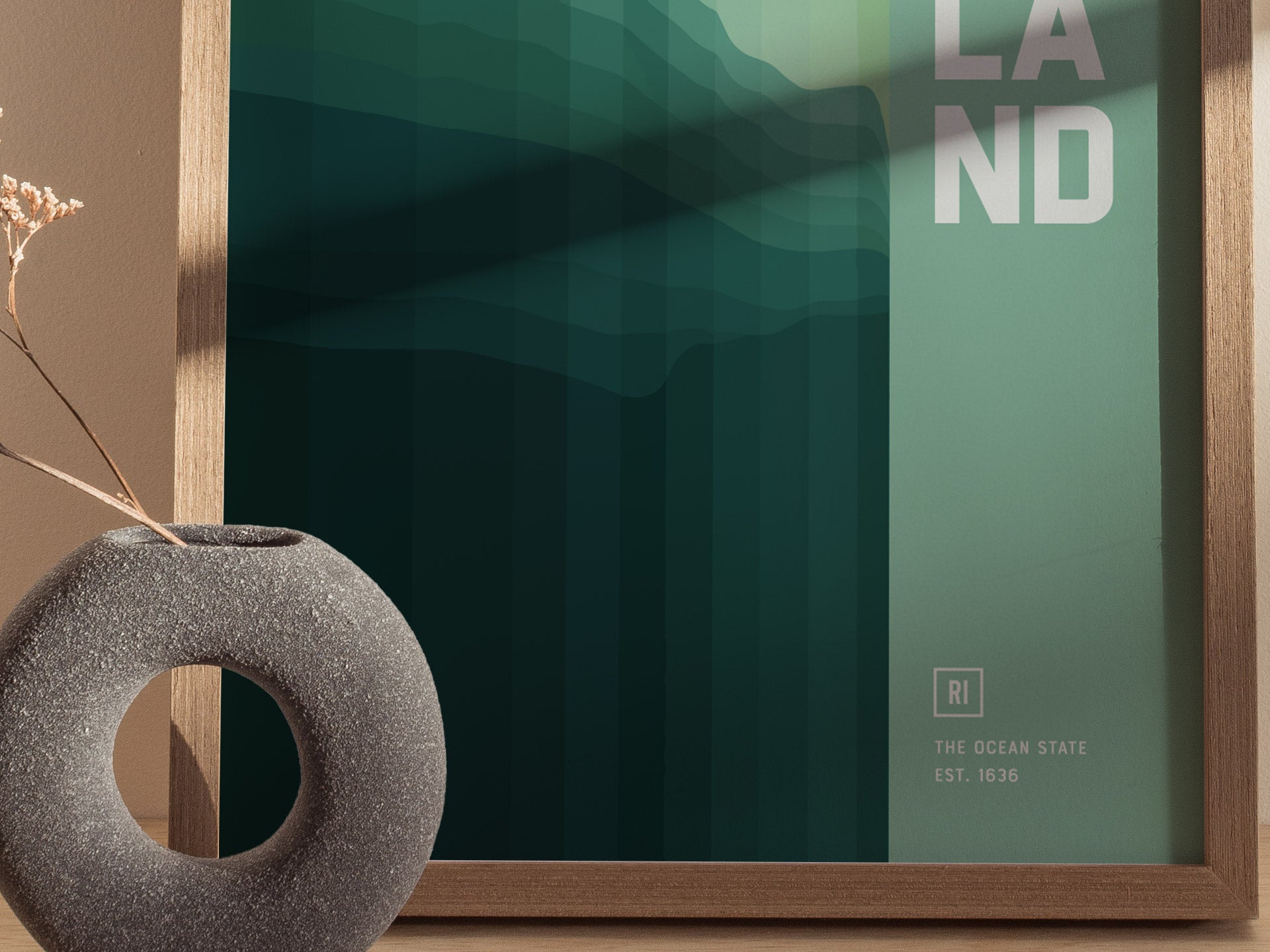 A framed abstract poster with green gradient waves and partial text LAND, beside a round, textured vase with dried flowers on a wooden surface.