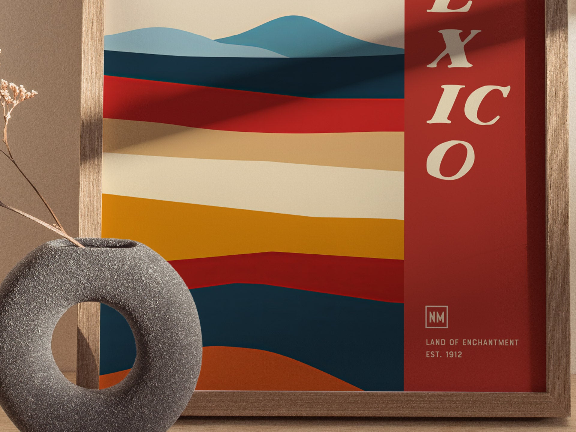 A colorful poster with abstract desert landscapes and bold red text reading EXICO, framed in wood. A textured, circular stone vase with dried flowers is in the foreground.
