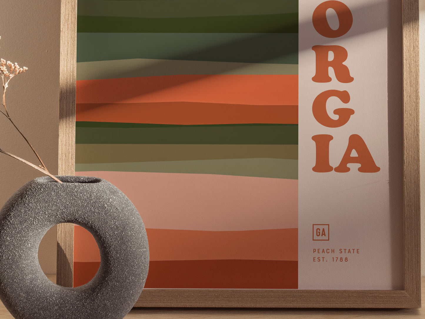 A framed poster with earthy, horizontal abstract lines and bold lettering spelling ORGIA is displayed beside a round, textured vase with dried flowers.