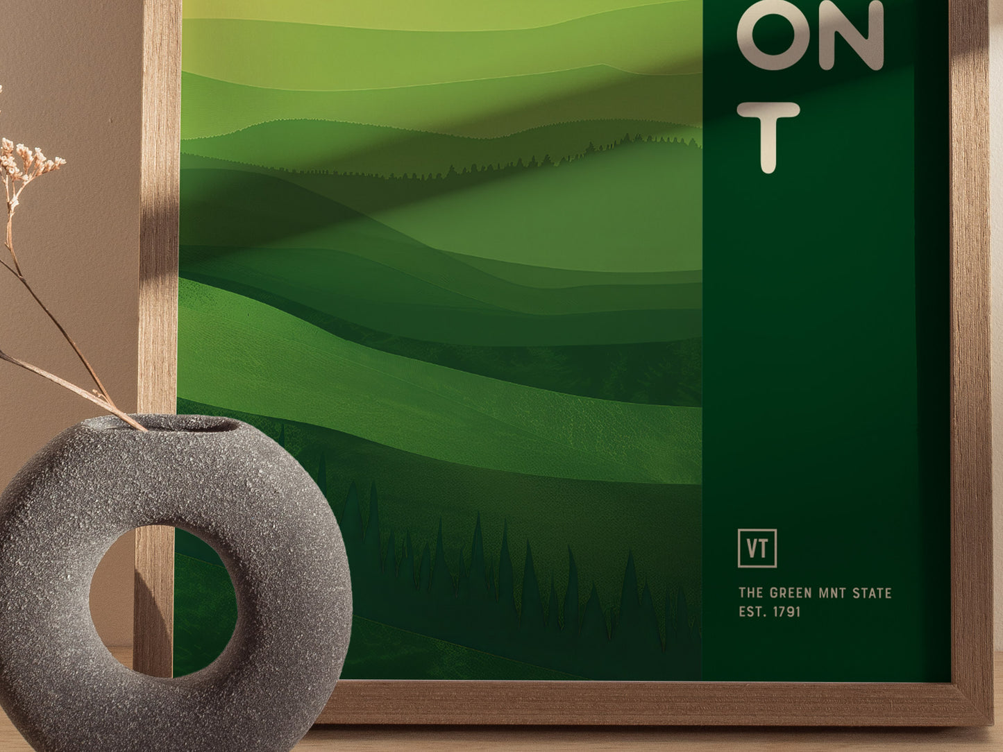 A framed poster with abstract green layers depicting rolling hills and forests, labeled VT The Green Mnt State Est. 1791, beside a round, gray vase with dried flowers.