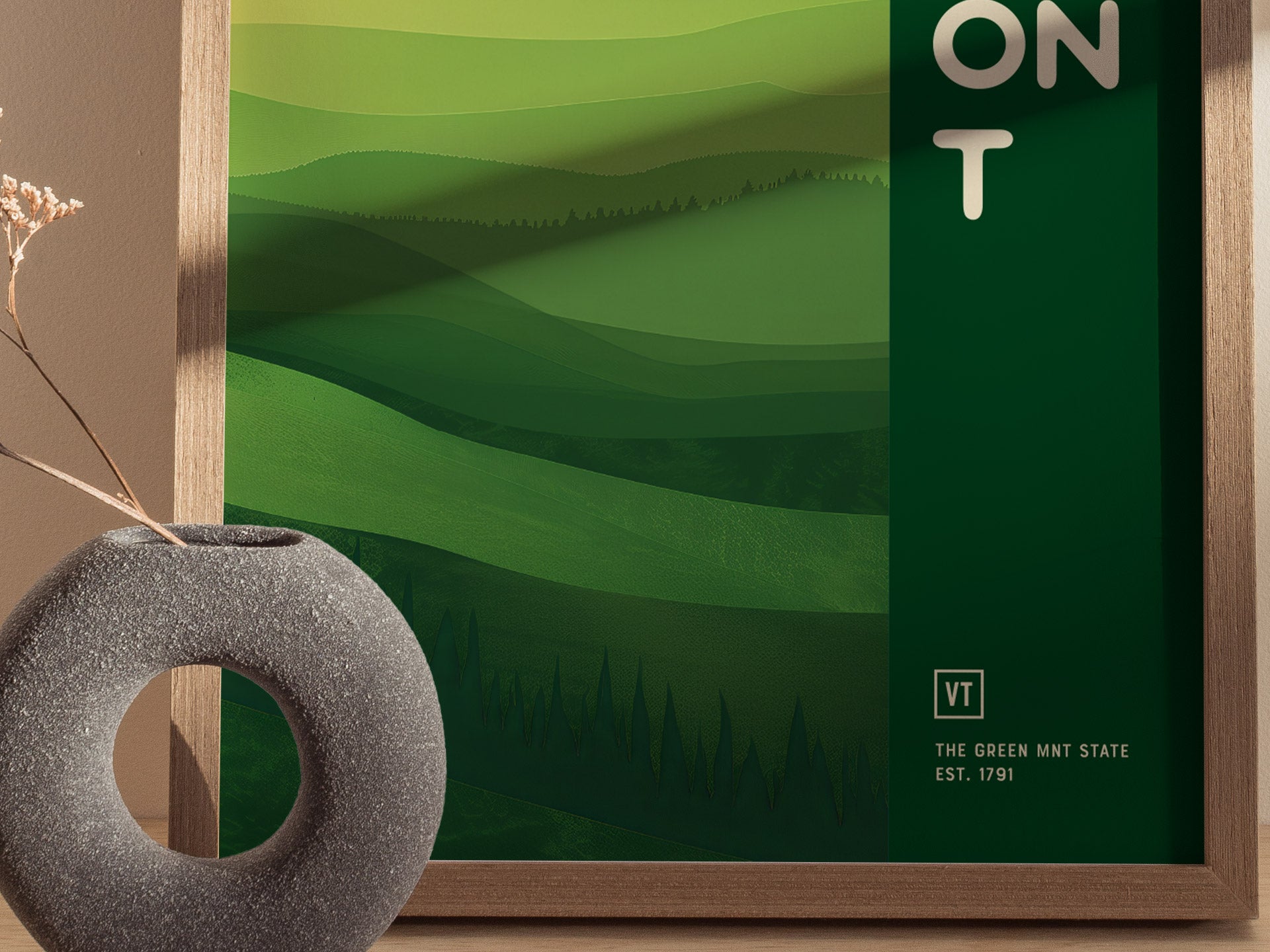 A framed poster with abstract green layers depicting rolling hills and forests, labeled VT The Green Mnt State Est. 1791, beside a round, gray vase with dried flowers.