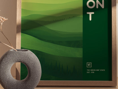 A framed poster with abstract green layers depicting rolling hills and forests, labeled VT The Green Mnt State Est. 1791, beside a round, gray vase with dried flowers.