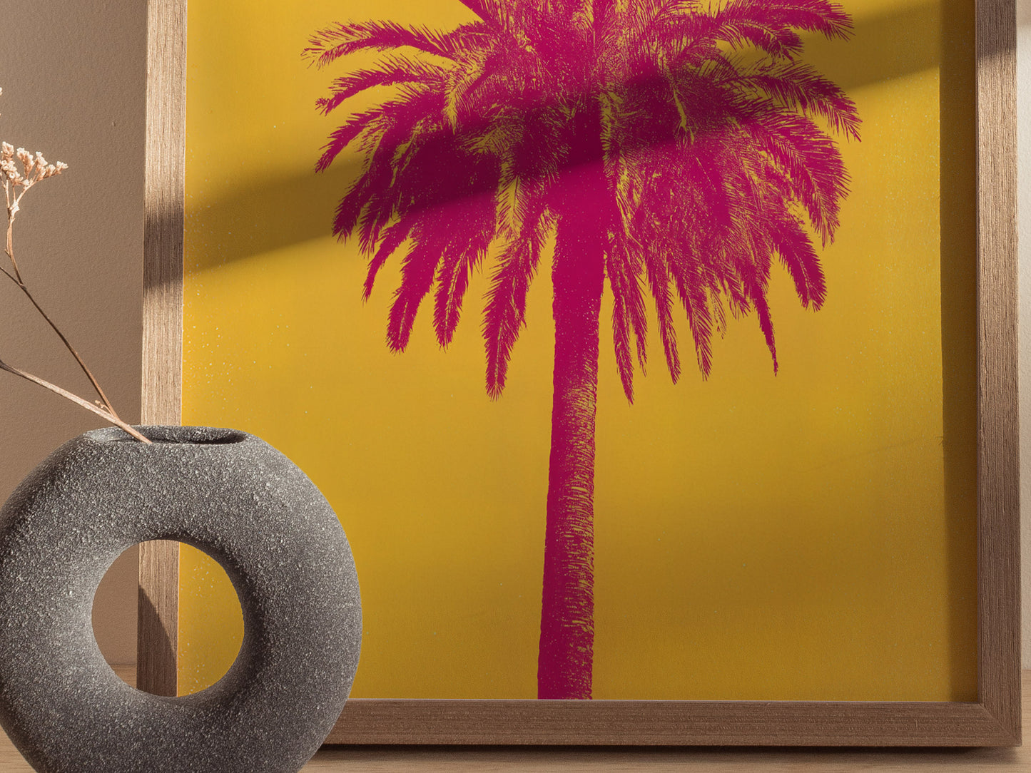 A vibrant poster features a pink palm tree against a yellow background, framed in wood. A round, textured gray vase with a dried plant is placed beside it.