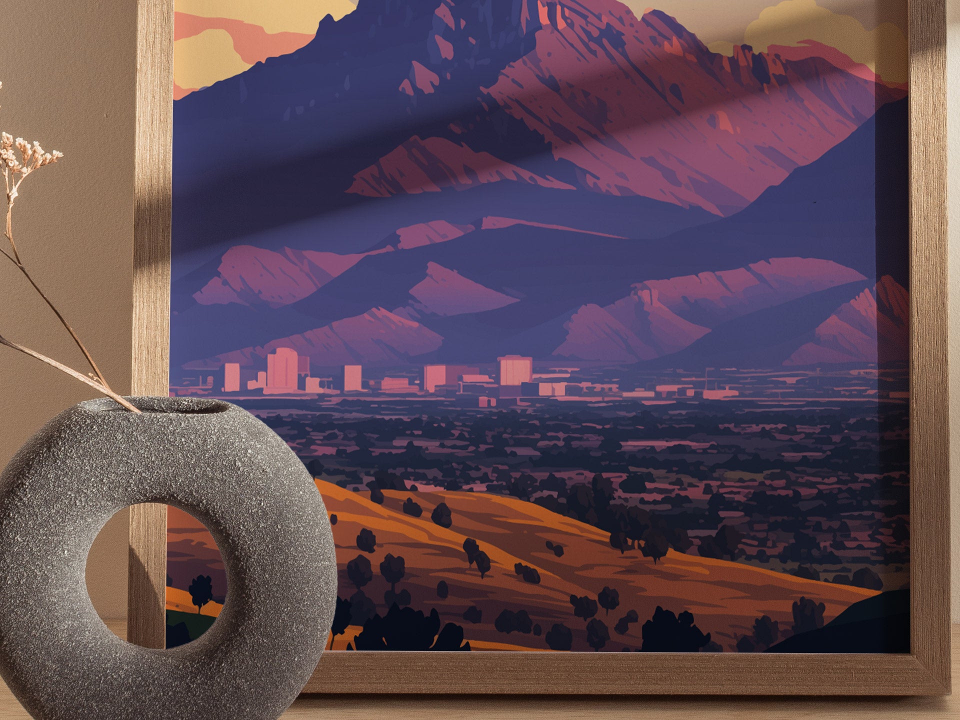 Framed artwork depicting a colorful landscape of Camelback Mountain with a cityscape below, set against a warm, earthy-toned wall and a textured, circular vase in the foreground.