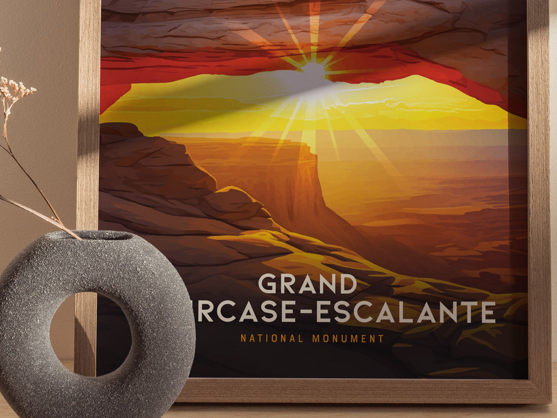 A framed poster depicts a vibrant sunset over the Grand Staircase–Escalante National Monument, with sun rays illuminating the red rock formations. A round, textured vase sits beside it.