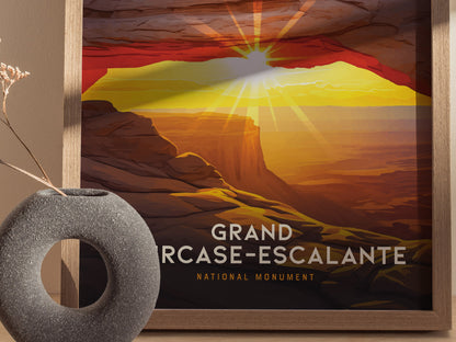 A framed poster depicts a vibrant sunset over the Grand Staircase–Escalante National Monument, with sun rays illuminating the red rock formations. A round, textured vase sits beside it.