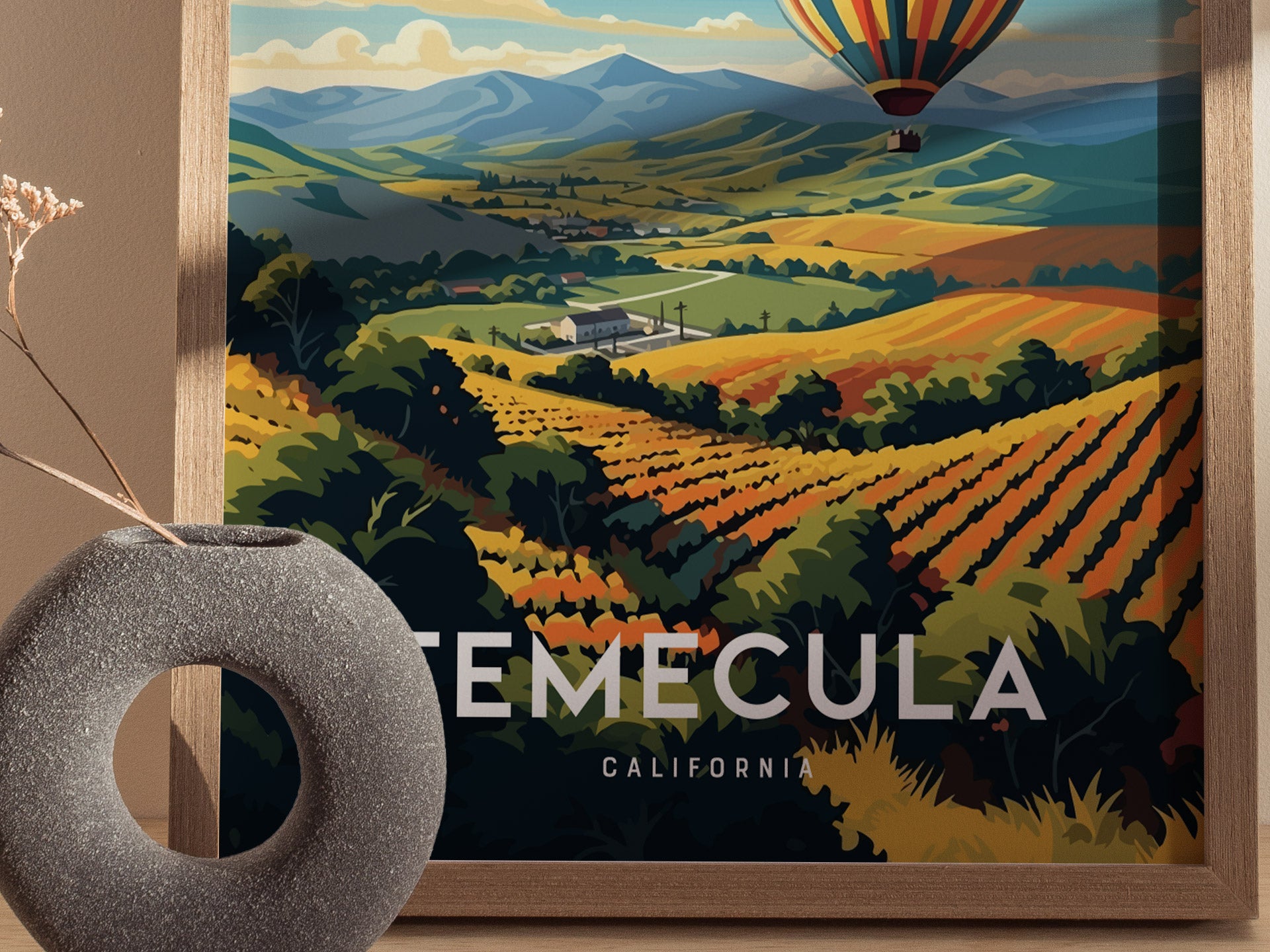 A framed poster featuring vibrant vineyards, rolling hills, and a colorful hot air balloon in Temecula, California. A gray vase with dried flowers is in the foreground.