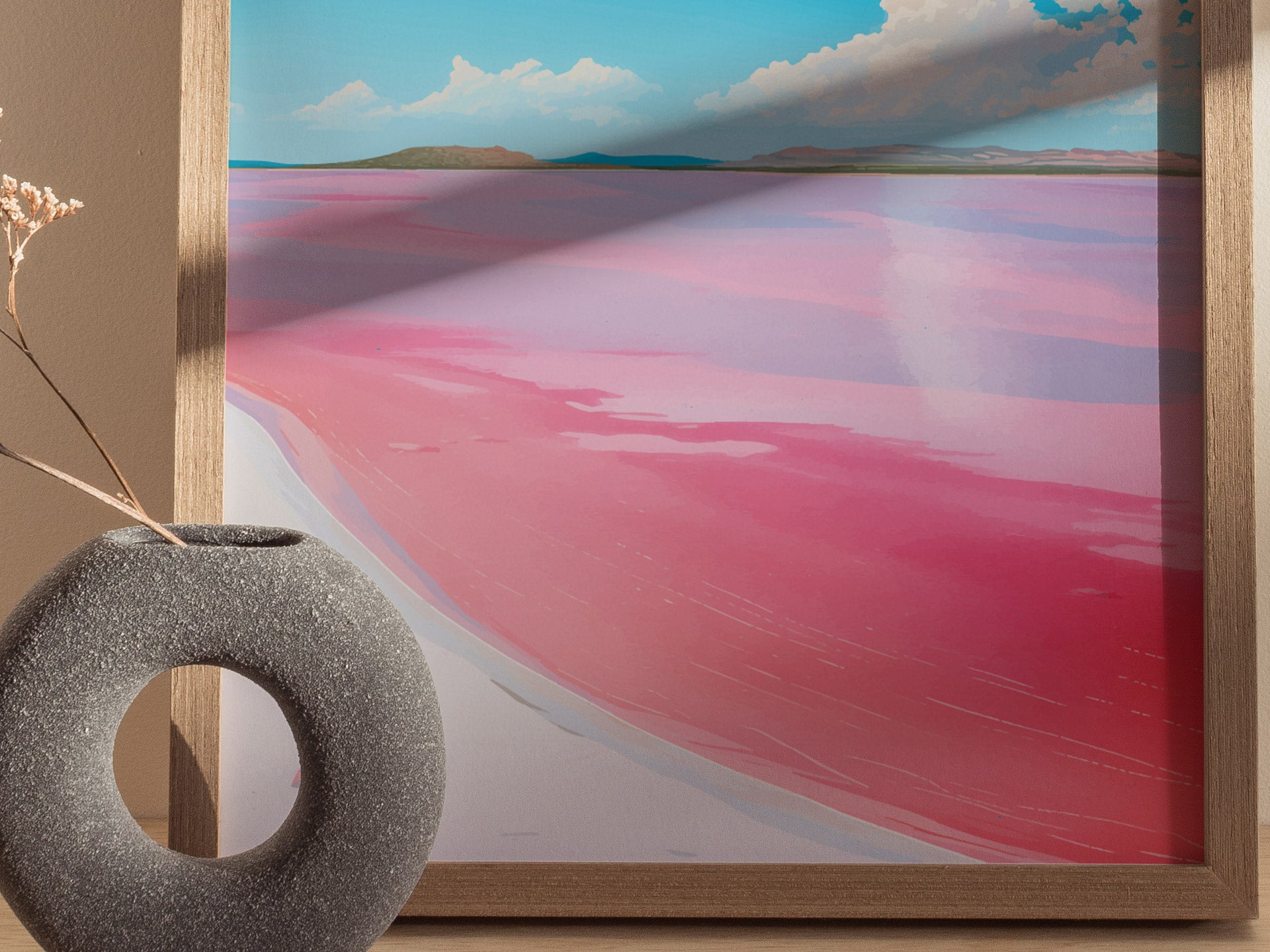 A framed poster depicts Lake Hillier with vibrant pink water contrasting against a blue sky, accompanied by a textured gray vase holding dry flowers in the foreground.