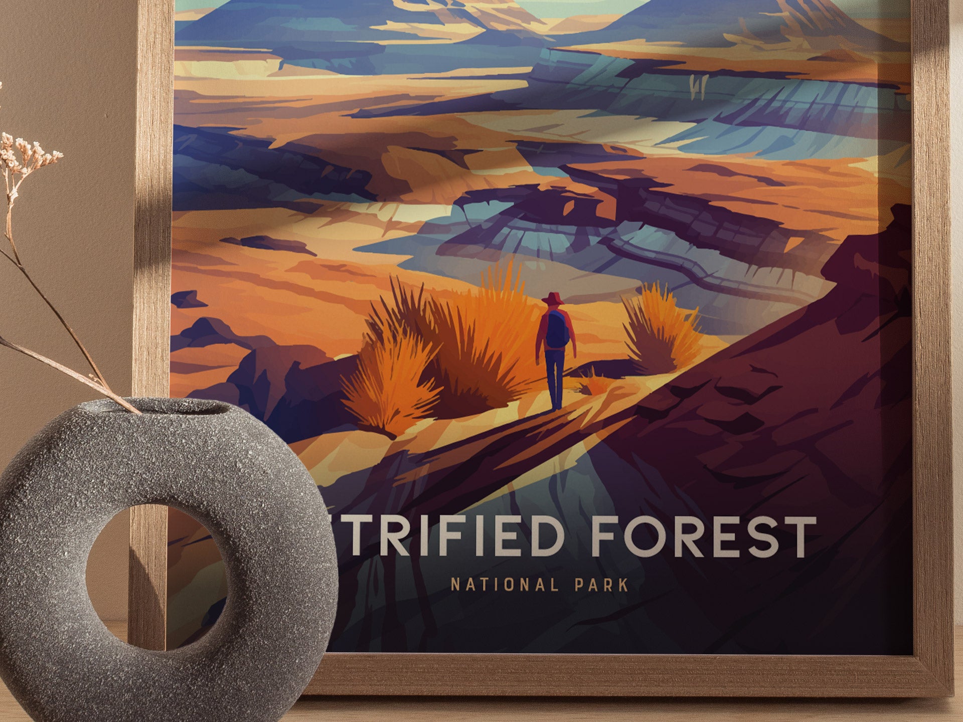 A colorful poster depicts a person walking through a vibrant desert landscape with layered rock formations and foliage. A gray, textured vase with dried branches sits in the foreground.