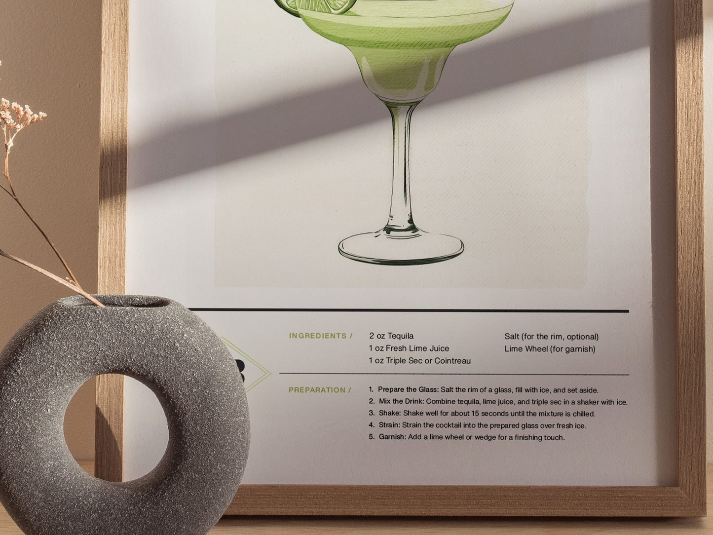 A framed poster of a margarita cocktail with ingredients and preparation instructions is displayed next to a round, textured vase containing dried flowers.