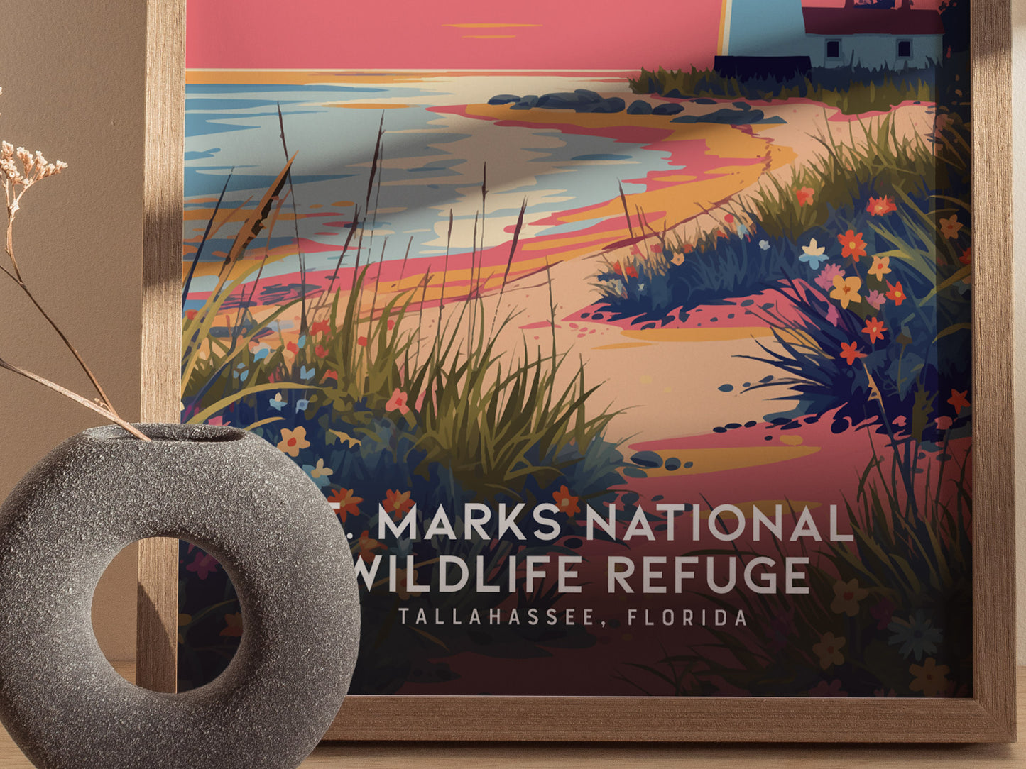 A framed poster depicts a colorful coastal scene with a lighthouse, vibrant wildflowers, and a sunset sky. A textured stone vase with dried branches is placed in front.
