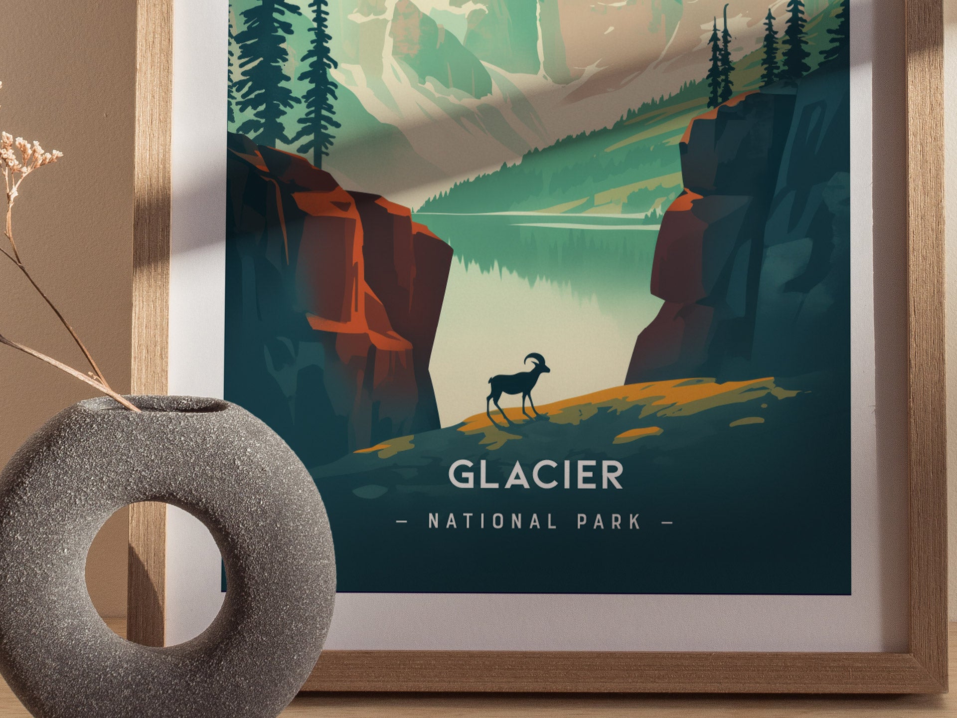 A framed minimalist poster of Glacier National Park features a bighorn sheep, alpine lake, and towering peaks. A textured vase with dried flowers is in the foreground.