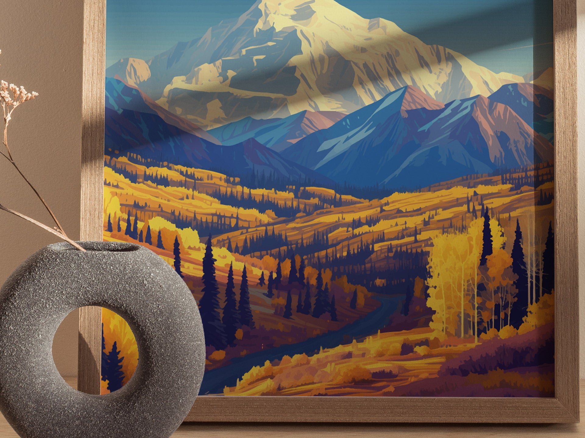 A framed poster depicting Mount Denali with vibrant yellow and orange fall foliage in the foreground. A gray, round vase with dried branches sits nearby.