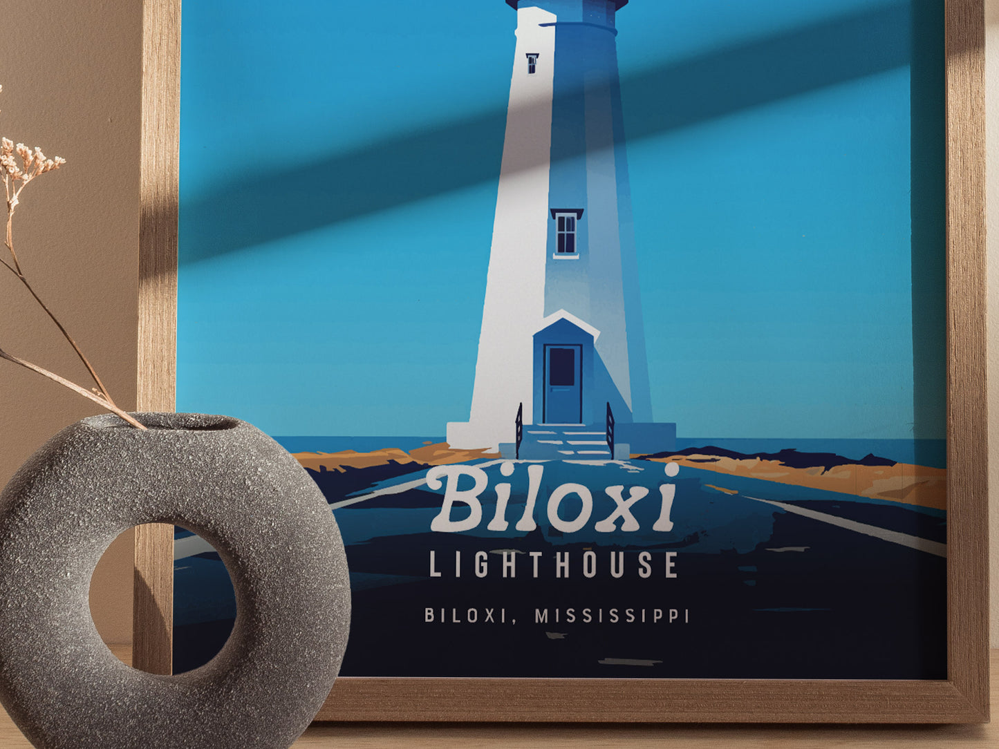 A framed poster depicts the Biloxi Lighthouse in a minimal vector style with a blue background. A textured gray vase with dried flowers is placed in the foreground.