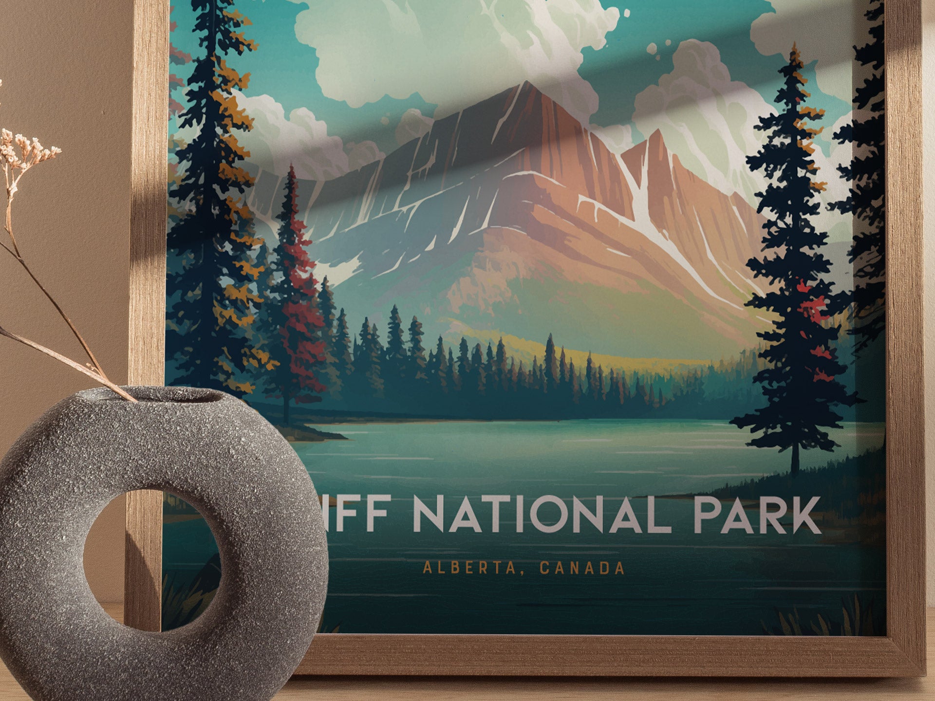 A framed poster of Banff National Park depicts majestic mountains and a serene lake with surrounding pine trees. A textured stone vase with dried flowers is in the foreground.
