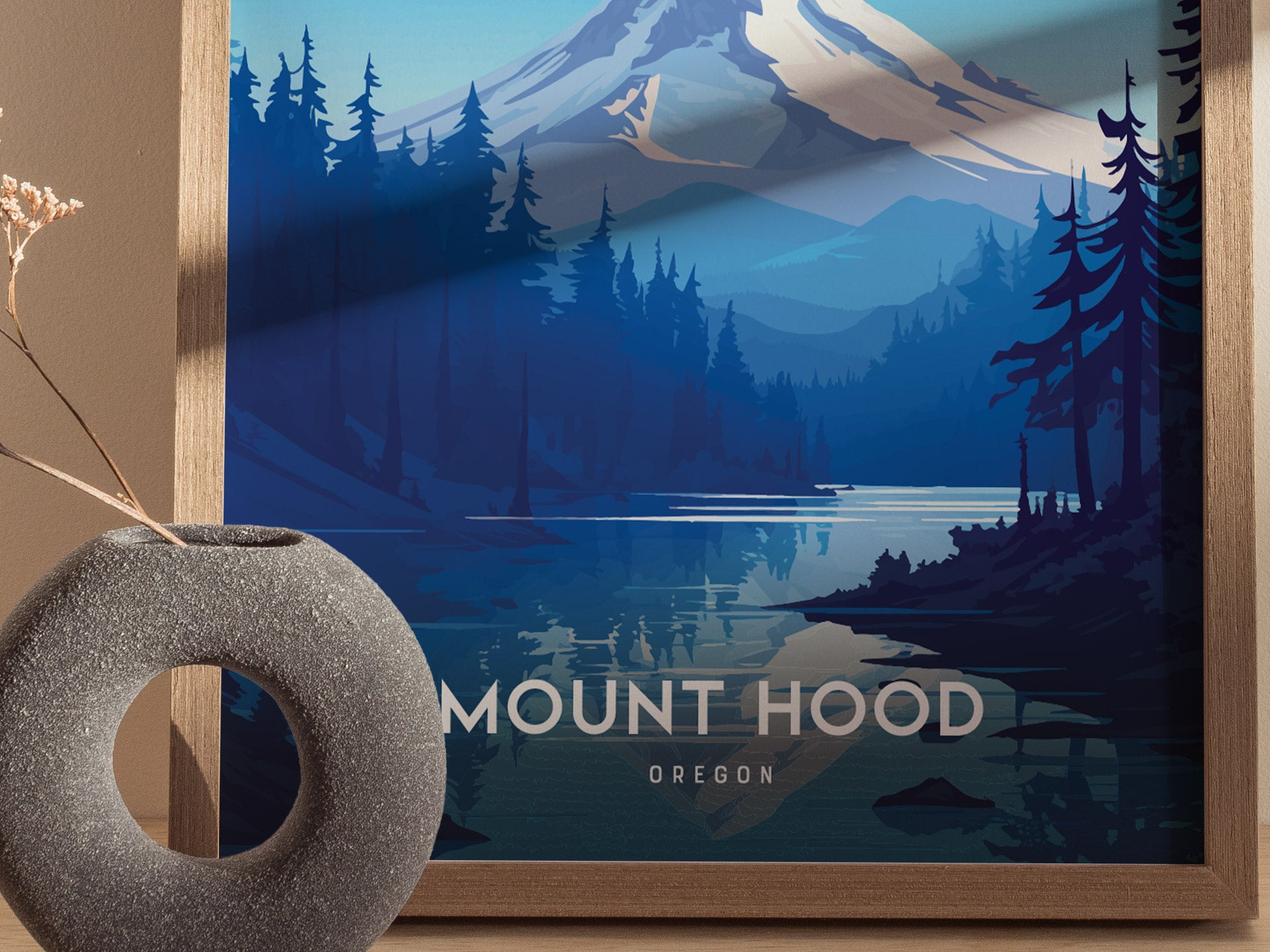 A framed Mount Hood poster features a stylized depiction of the mountain with forested foothills and a serene lake. A textured gray vase sits nearby, enhancing the composition.