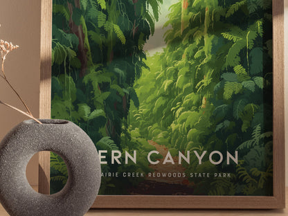A framed poster of Fern Canyon features lush green foliage with a path leading through the dense vegetation. A round, textured vase sits in the foreground.