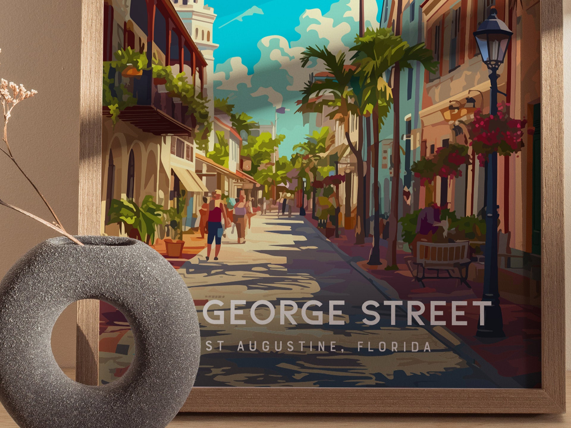 A colorful poster of St. George Street in St. Augustine, Florida, displays vibrant buildings, palm trees, and pedestrians. A round, textured vase with dried flowers is in the foreground.