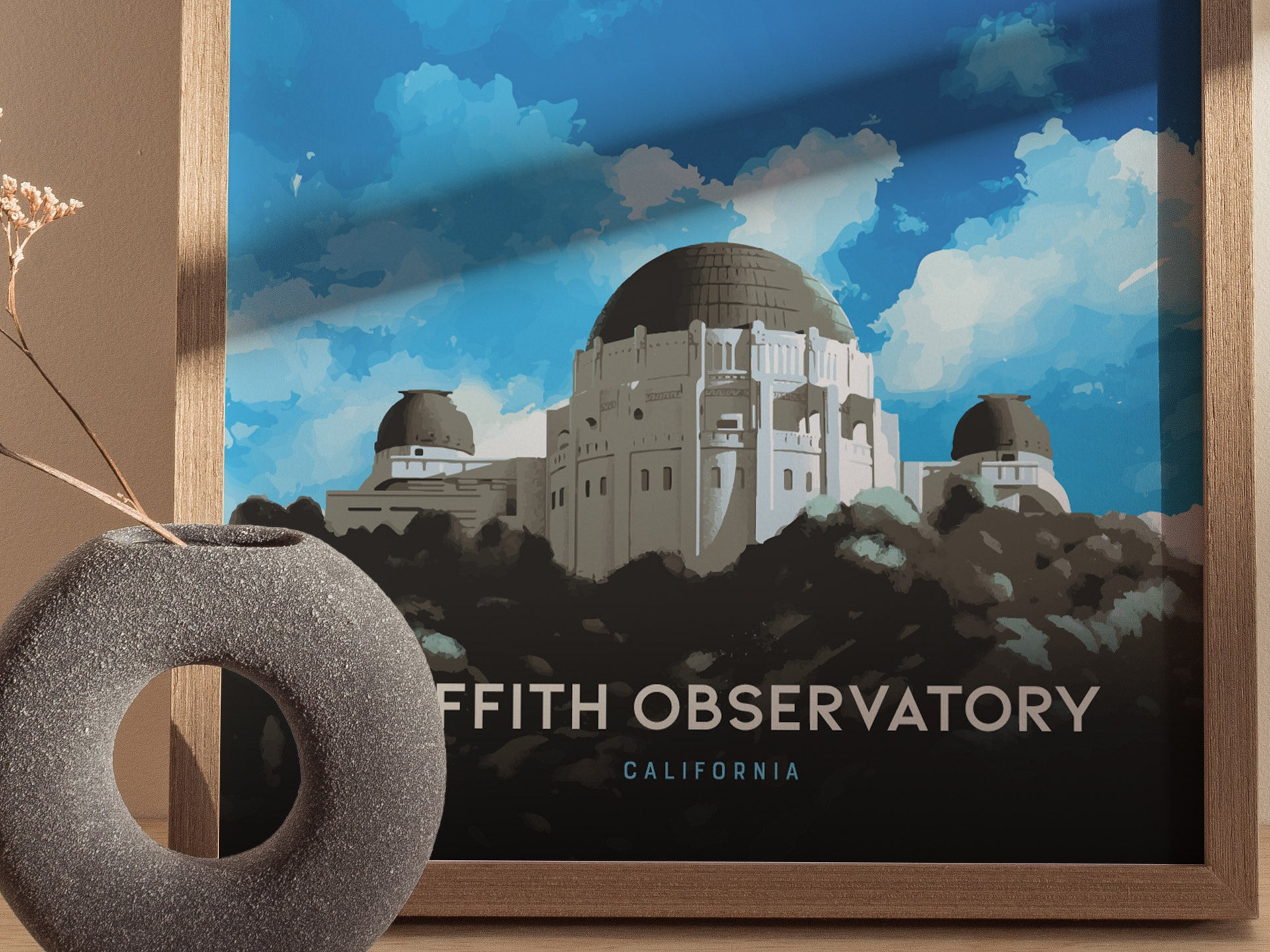 A framed poster of Griffith Observatory features its iconic domes against a vibrant blue sky. In the foreground, a round, textured vase holds dried flowers.