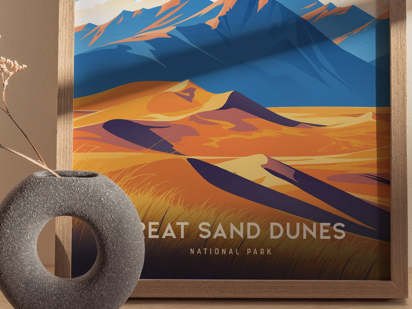 A framed poster featuring stylized, colorful illustrations of sand dunes and mountains, labeled Great Sand Dunes National Park, sits beside a textured, circular vase on a wooden surface.