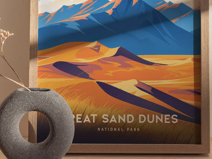 A framed poster featuring stylized, colorful illustrations of sand dunes and mountains, labeled Great Sand Dunes National Park, sits beside a textured, circular vase on a wooden surface.