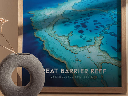A framed poster of the Great Barrier Reef with vibrant blue and turquoise waters is displayed next to a gray, circular vase on a wooden surface.