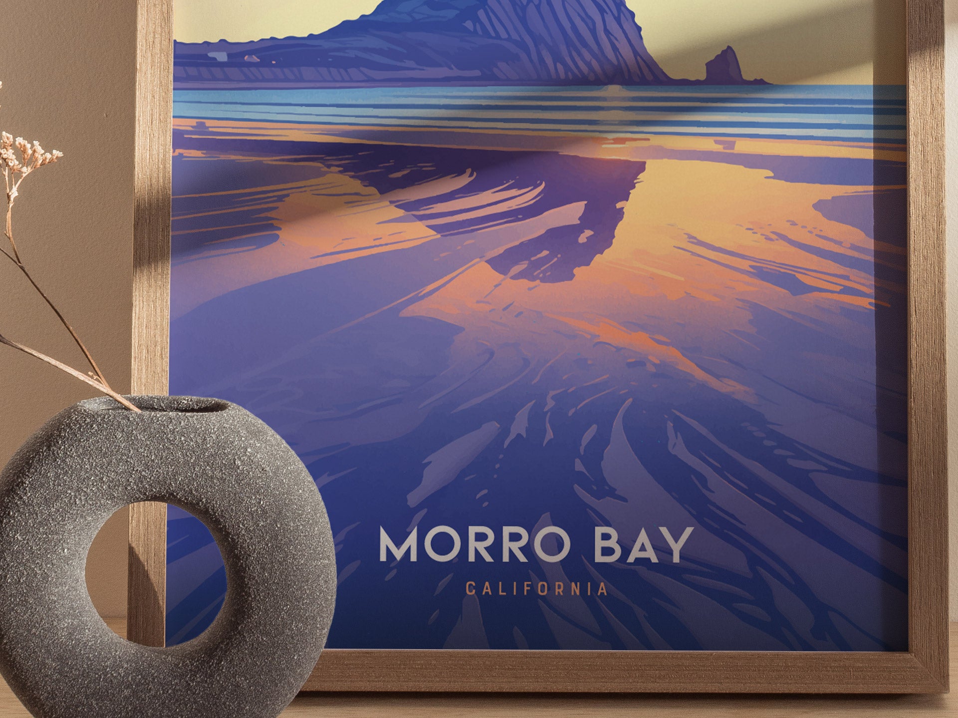 A framed Morro Bay poster showcases a stylized coastal landscape with vibrant hues of blue and orange. In front, a textured round vase holds a dried plant.