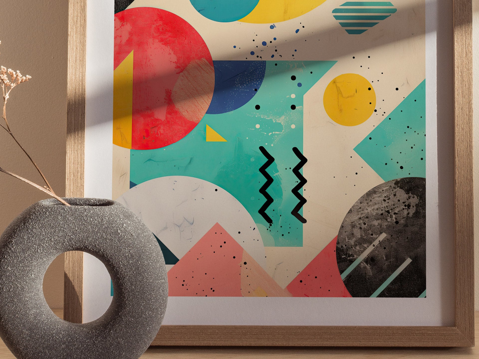 A framed poster featuring vibrant 90s-inspired geometric shapes and colors, including circles, triangles, and zigzag lines, is displayed next to a textured gray vase with dried flowers.