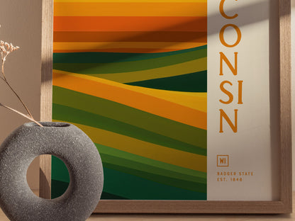 A framed abstract poster features layered green and gold waves with the word Wisconsin vertically on the side. A textured gray vase with dried flowers is in the foreground.