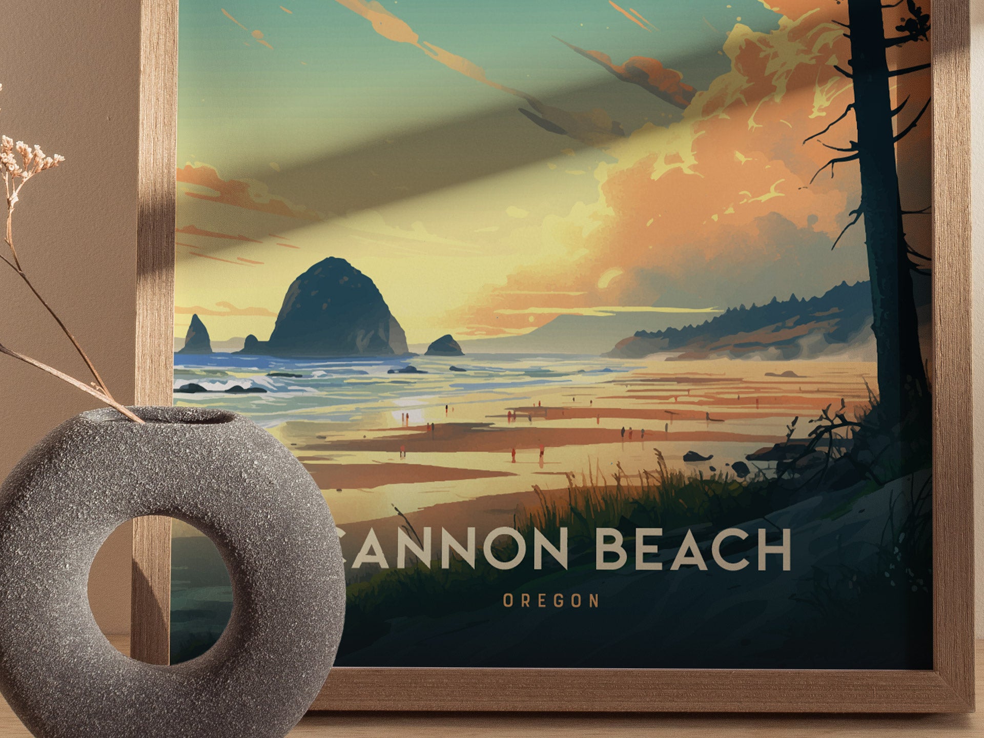 A framed poster depicting Cannon Beach, Oregon, showcases a sunset over Haystack Rock with people on the sandy shore. A textured gray vase with dried flowers is in the foreground.