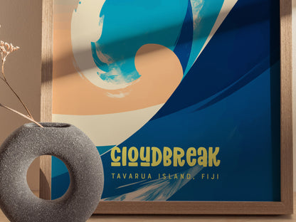 A vibrant surf poster with abstract wave designs in blue and beige, labeled Cloudbreak Tavarua Island, Fiji, displayed in a wooden frame beside a textured round vase.