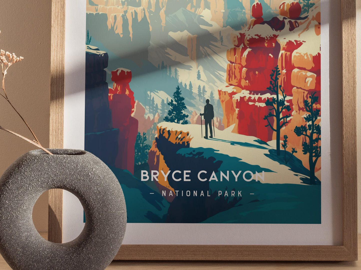 A framed Bryce Canyon National Park print features a hiker amid vibrant red rock formations and snowy landscapes. A textured round vase with dried flowers sits in front of it.