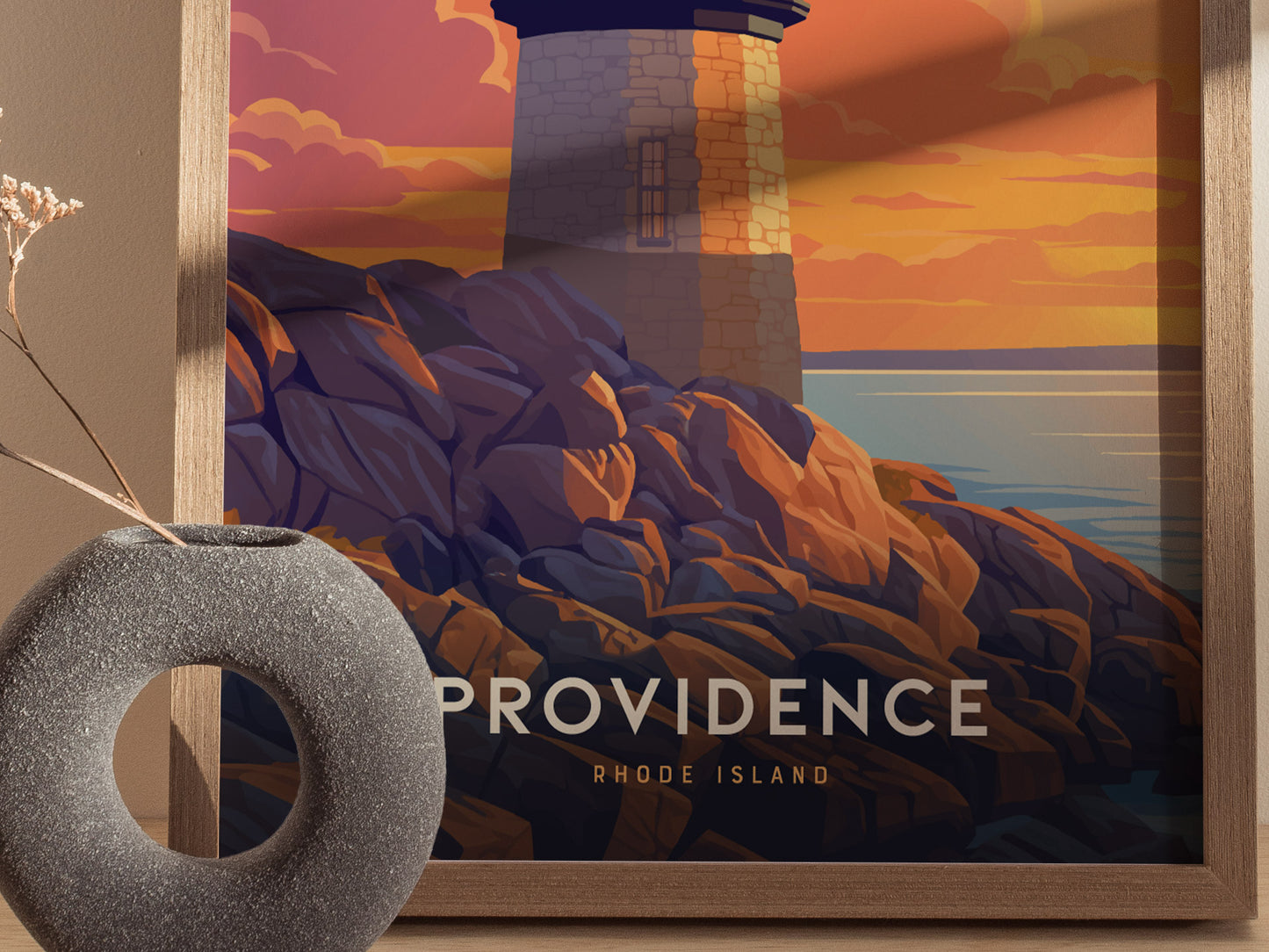 A framed poster depicts a lighthouse on rocky cliffs at sunset, with bold text reading Providence Rhode Island. A gray circular vase is placed nearby.