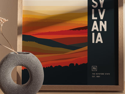 A framed abstract poster featuring vibrant orange and red layered landscapes with the word SYLVANIA in bold letters. A textured circular vase with dried flowers sits nearby.