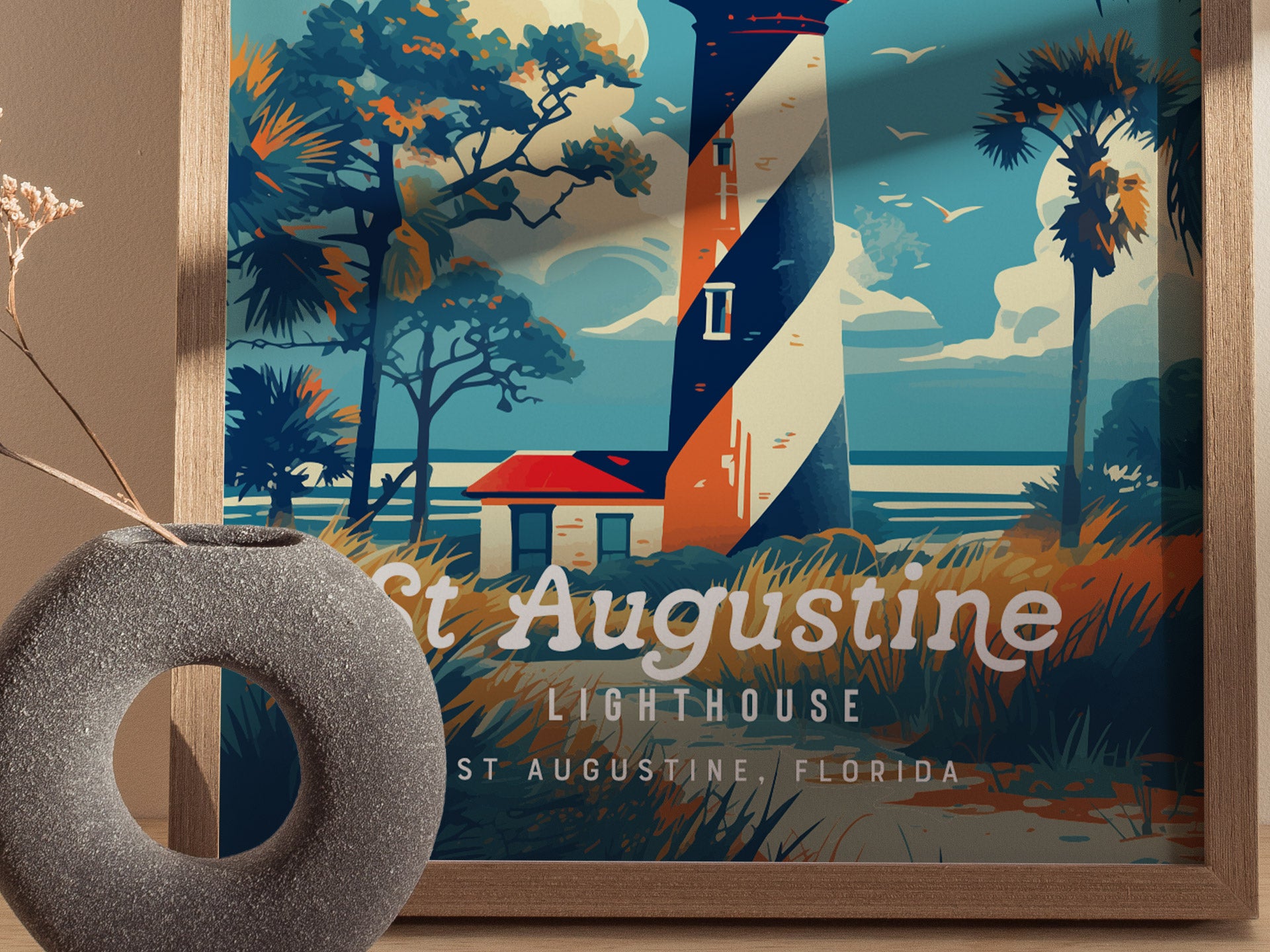 A framed poster of the St. Augustine Lighthouse featuring a black and white spiral tower set against a vibrant coastal backdrop with palm trees and a red-roofed building. A decorative round vase is in the foreground.