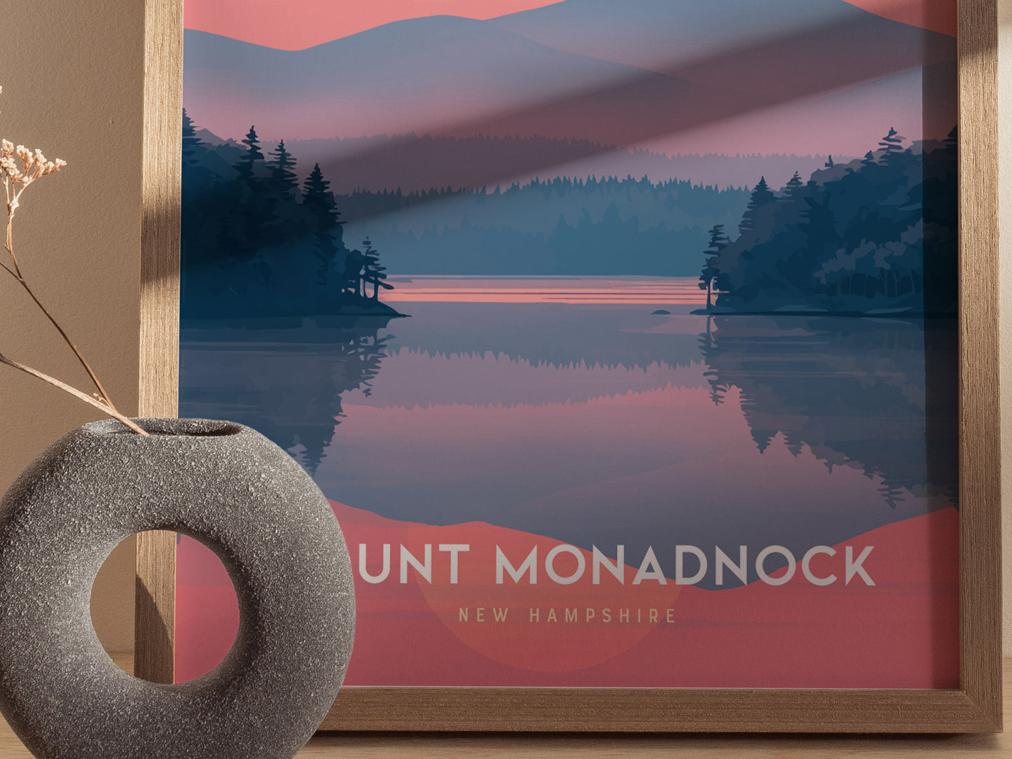 Framed poster of Mount Monadnock in pink and blue hues, depicting a serene lake and forest landscape. A round, textured vase with dried flowers is in the foreground.