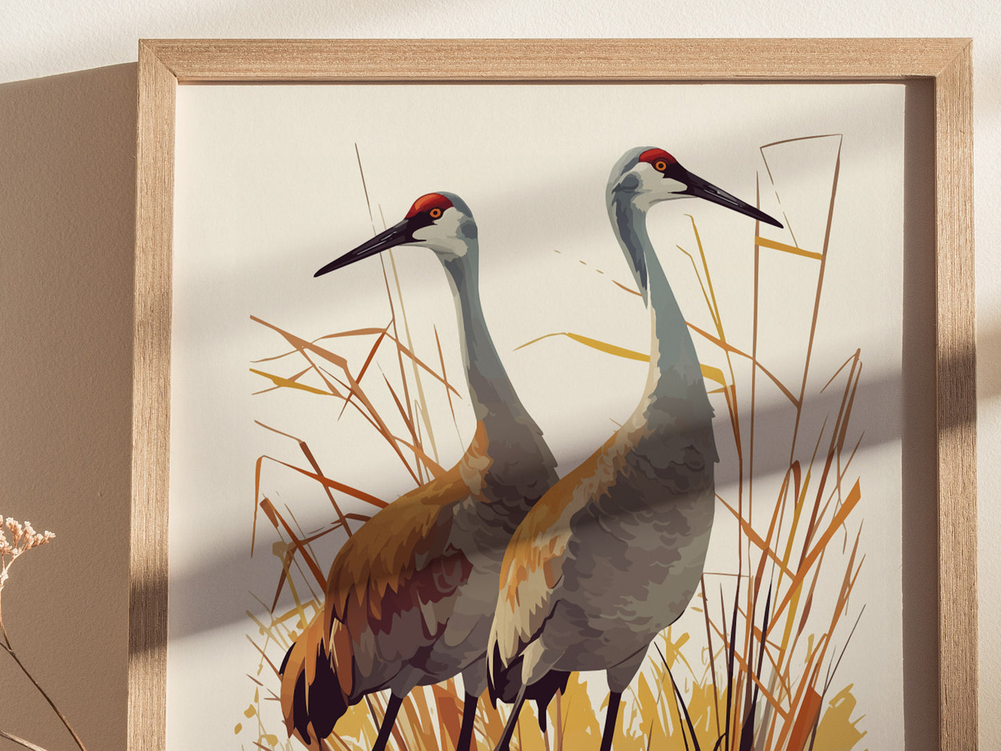 A framed poster depicts two sandhill cranes standing among tall grasses, rendered in soft browns and grays with a minimalist style. Shadows cast on the wall add depth.